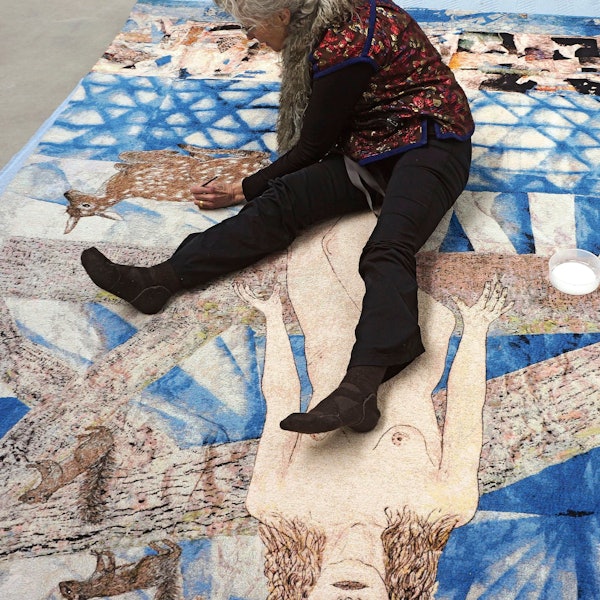 Kiki Smith sitzt auf einer über den Boden ausgebreiteten Papiercollage und malt mit einem Stift.