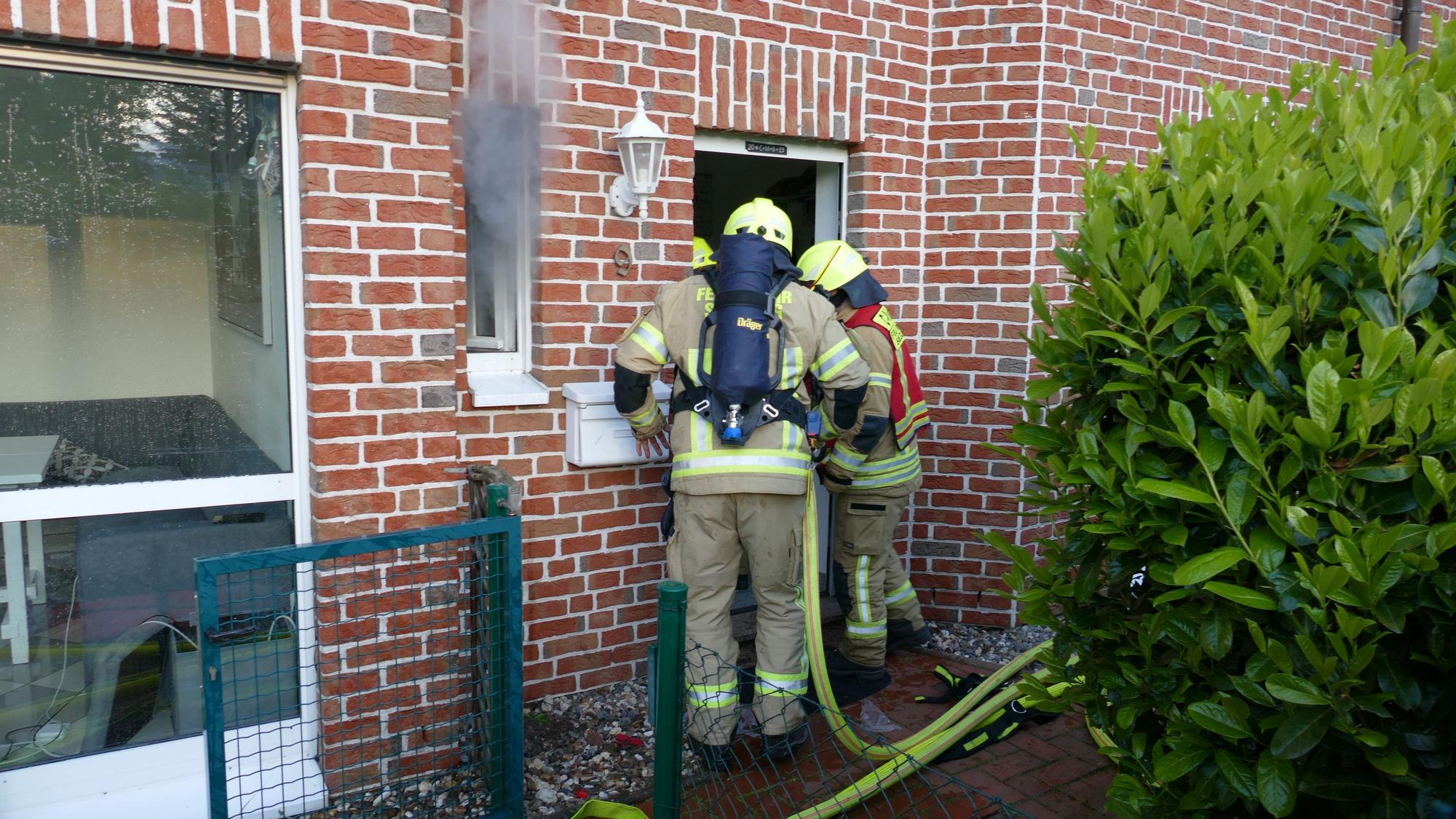 Feuerwehrleute stehen vor einer Haustür, aus dem Fenster daneben kommt Qualm.