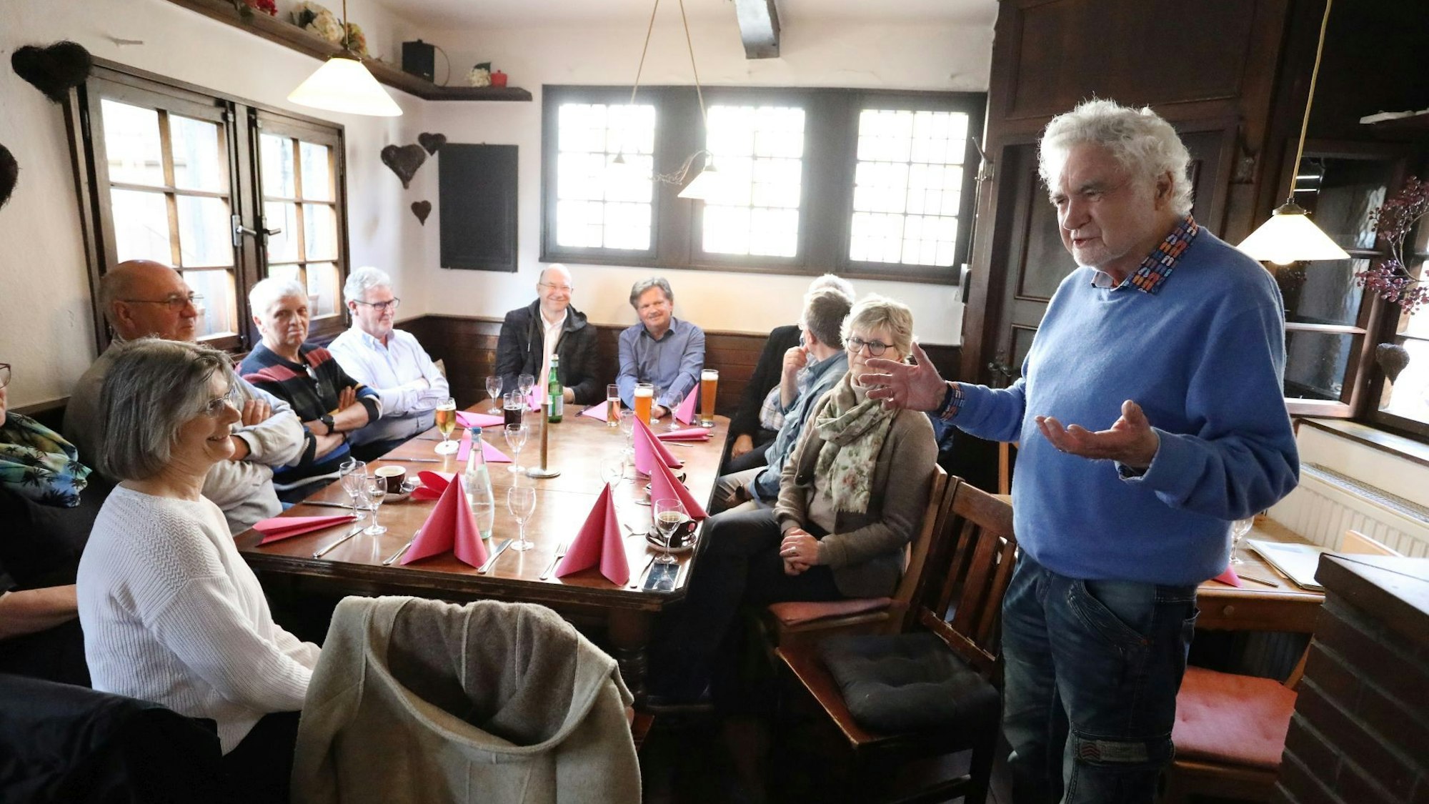 Ein Mann mit weißen Haaren und einem blauen Pullover spricht vor Zuhörern in einer Gaststätte.