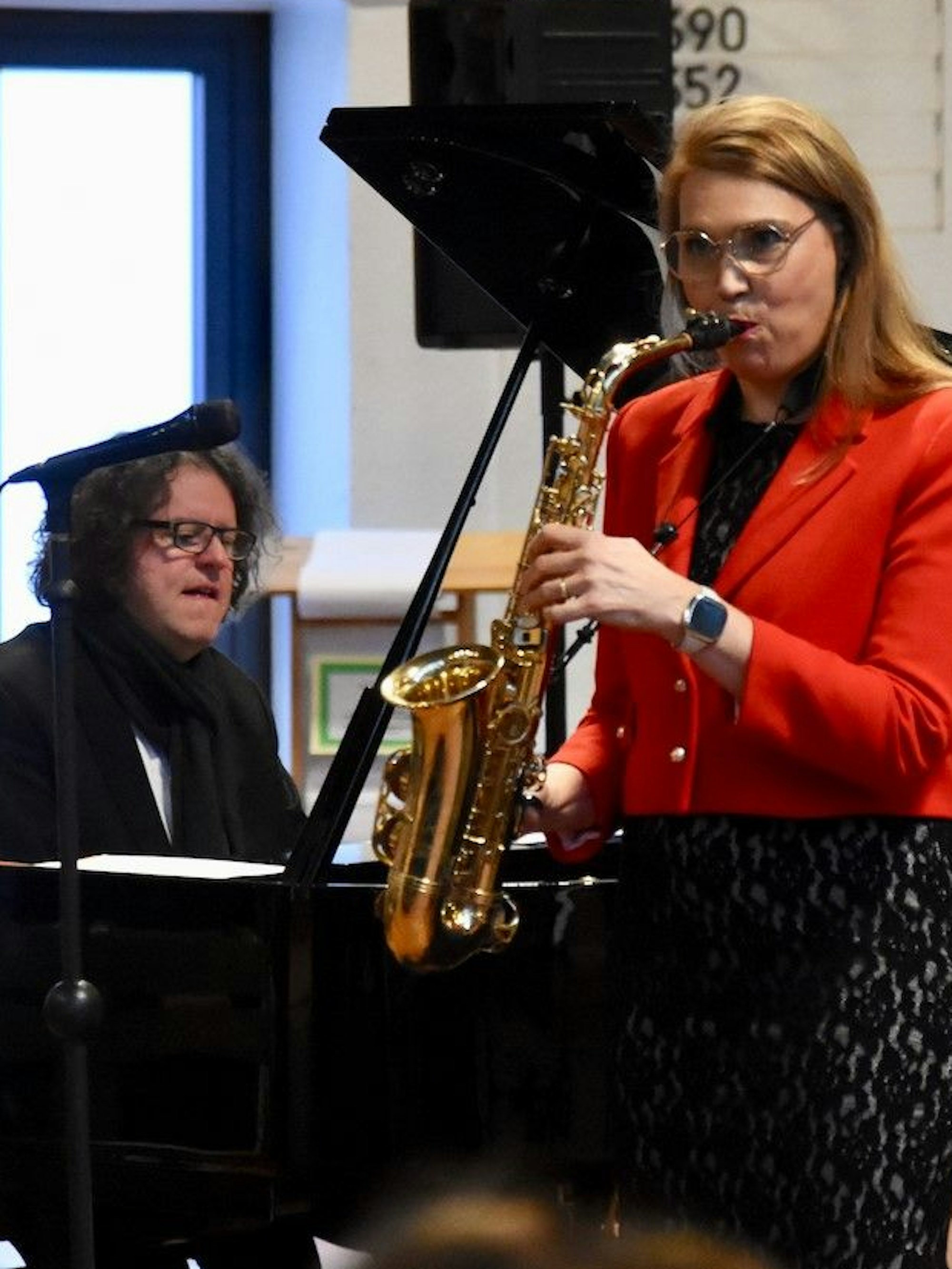 Eine Frau im roten Sakko spielt Saxophon, ein Mann mit Brille und lockigen Haaren begleitet sie am Flügel.