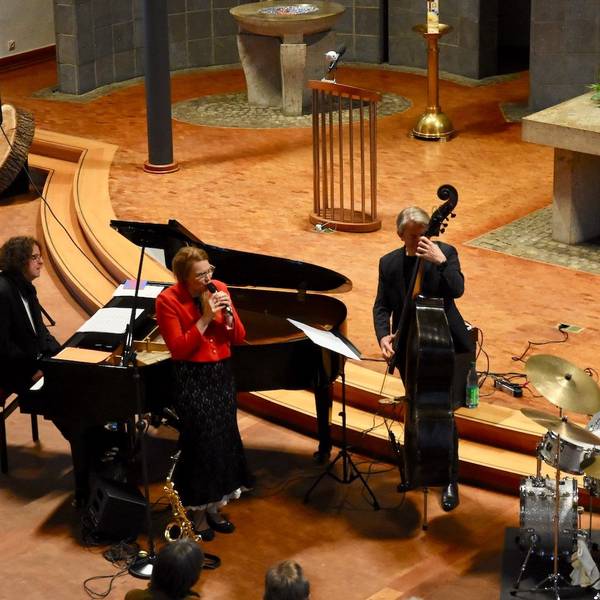 Männer und Frauen musizieren in einer Kirche. Sie spielen Bass, Schlagzeug und Klavier; eine Frau in einer roten Jacke singt.