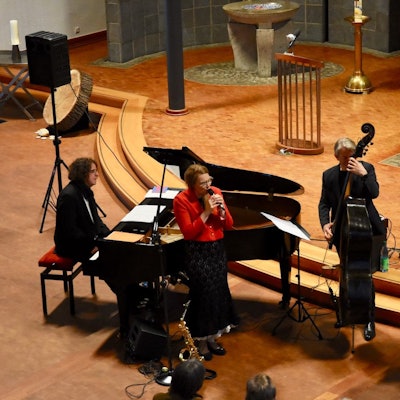 Männer und Frauen musizieren in einer Kirche. Sie spielen Bass, Schlagzeug und Klavier; eine Frau in einer roten Jacke singt.