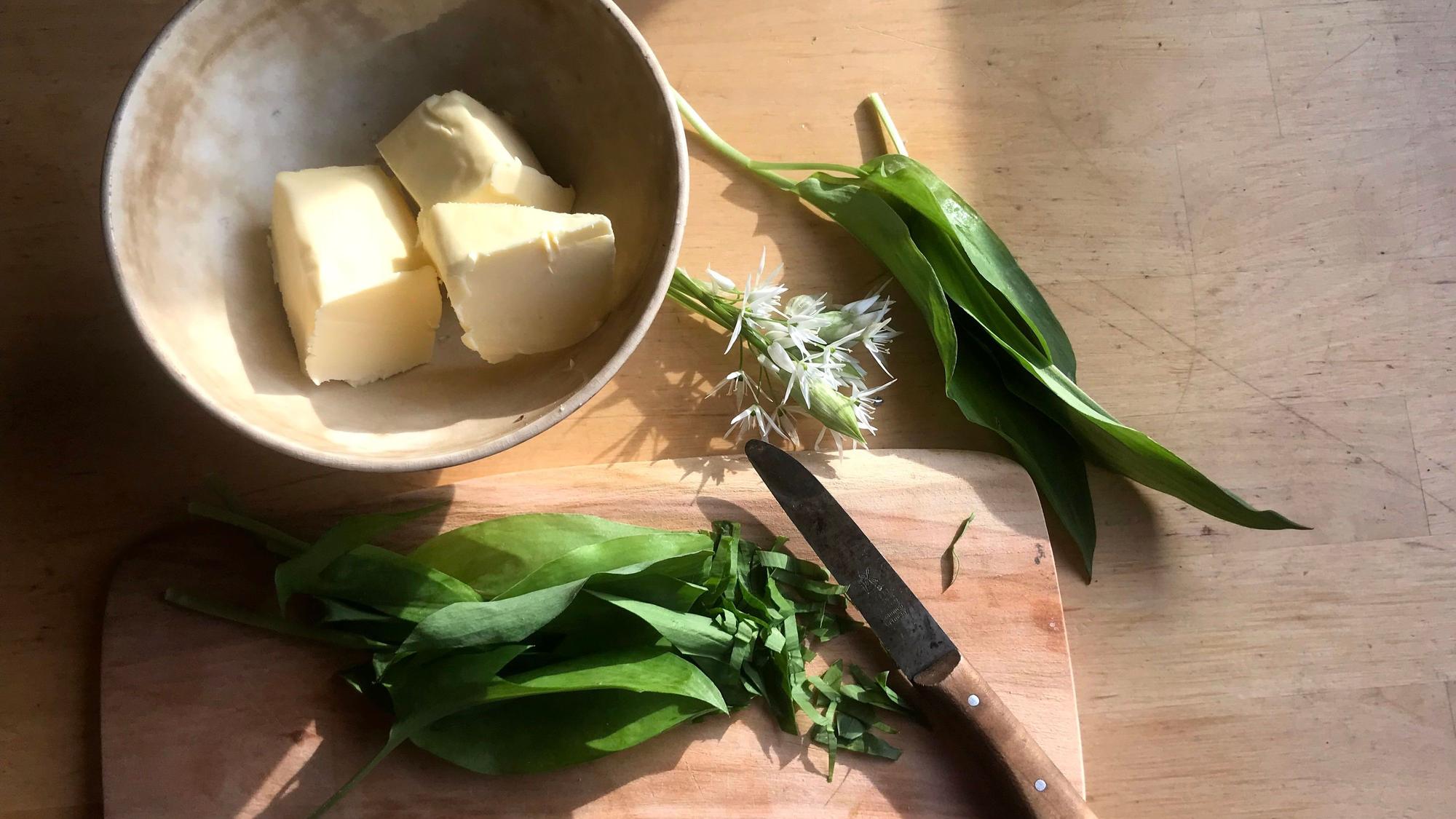 Drei Butterstücke in einer Tonschale, einige Bärlauchblätter und -blüten mit einem Messer auf einem Schneidebrett aus Holz.
