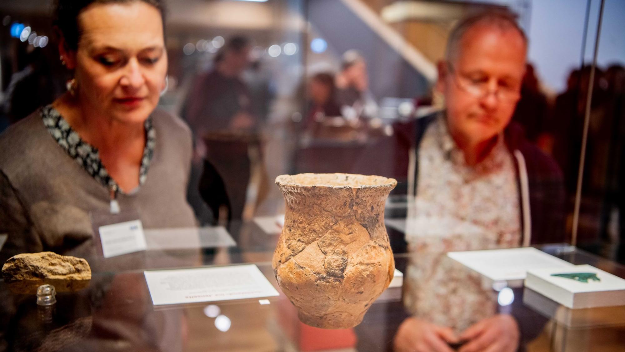 Das Bild zeigt einen Krug in einer Vitrine. Das Gefäß ist bei Großbüllesheim gefunden worden. Zwei Besucher schauen sich den Fund an.