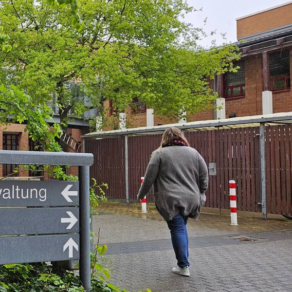 Das Bild zeigt ein Gebäude aus Backstein mit einem gläsernen Gang und einem grauen Schild davor.