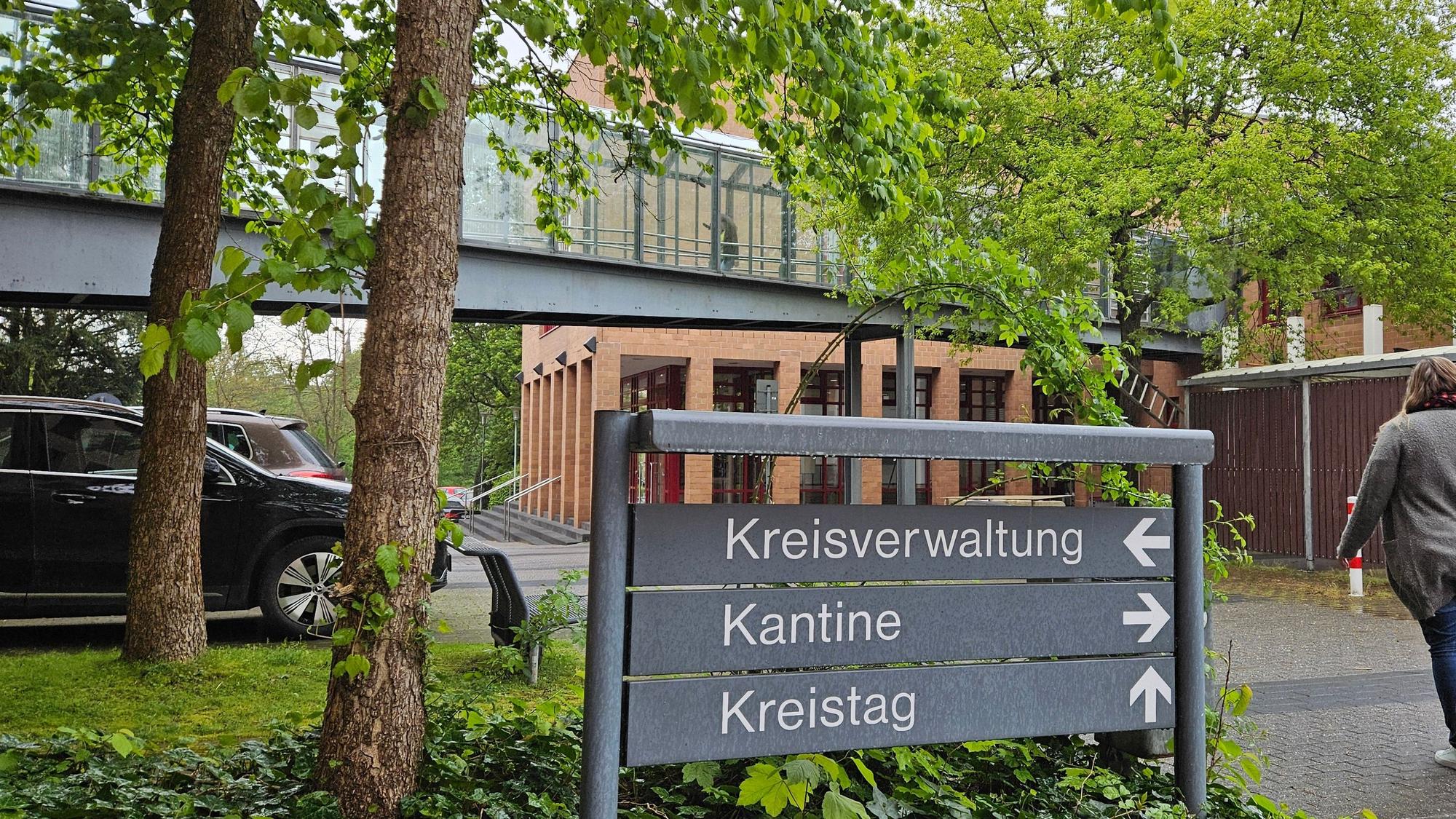 Das Bild zeigt ein Gebäude aus Backstein mit einem gläsernen Gang und einem grauen Schild davor.