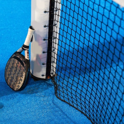 Das Bild zeigt einen Padel-Schläger, der an dem Pfosten eines Tennisnetzes lehnt. Auf dem blauen Court liegen zwei gelbe Tennisbälle.