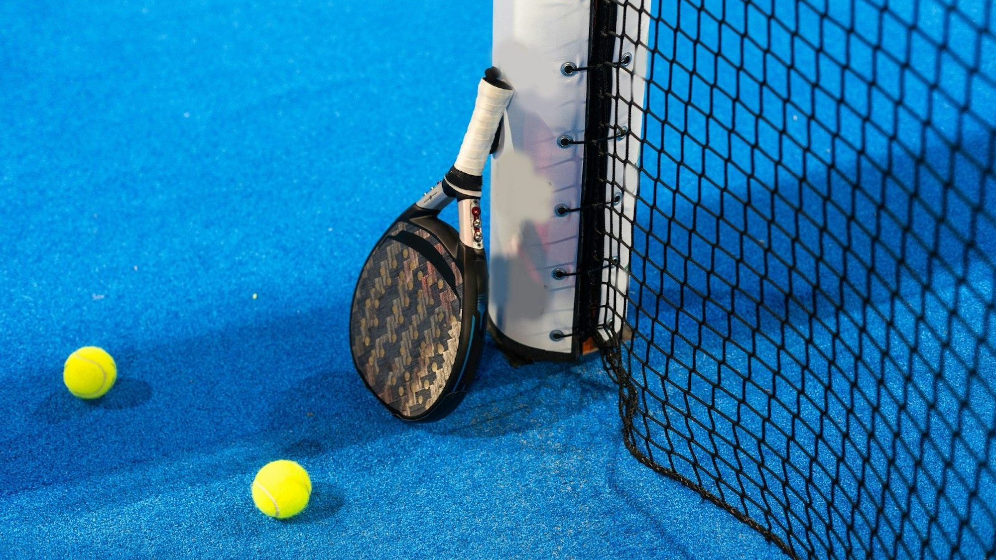 Das Bild zeigt einen Padel-Schläger, der an dem Pfosten eines Tennisnetzes lehnt. Auf dem blauen Court liegen zwei gelbe Tennisbälle.