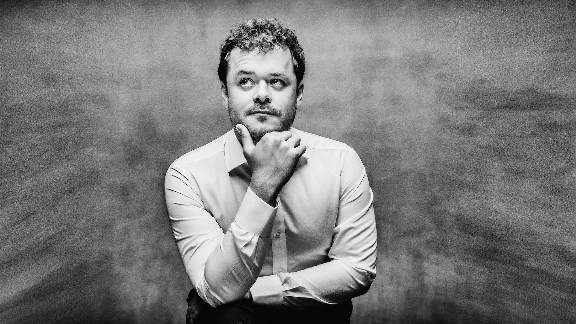 Das schwarz-weiße Bild zeigt Benjamin Grosvenor in nachdenklicher Pose, das Kinn stützt er mit der Hand. Der Hintergrund wirkt rauchig-verworren.