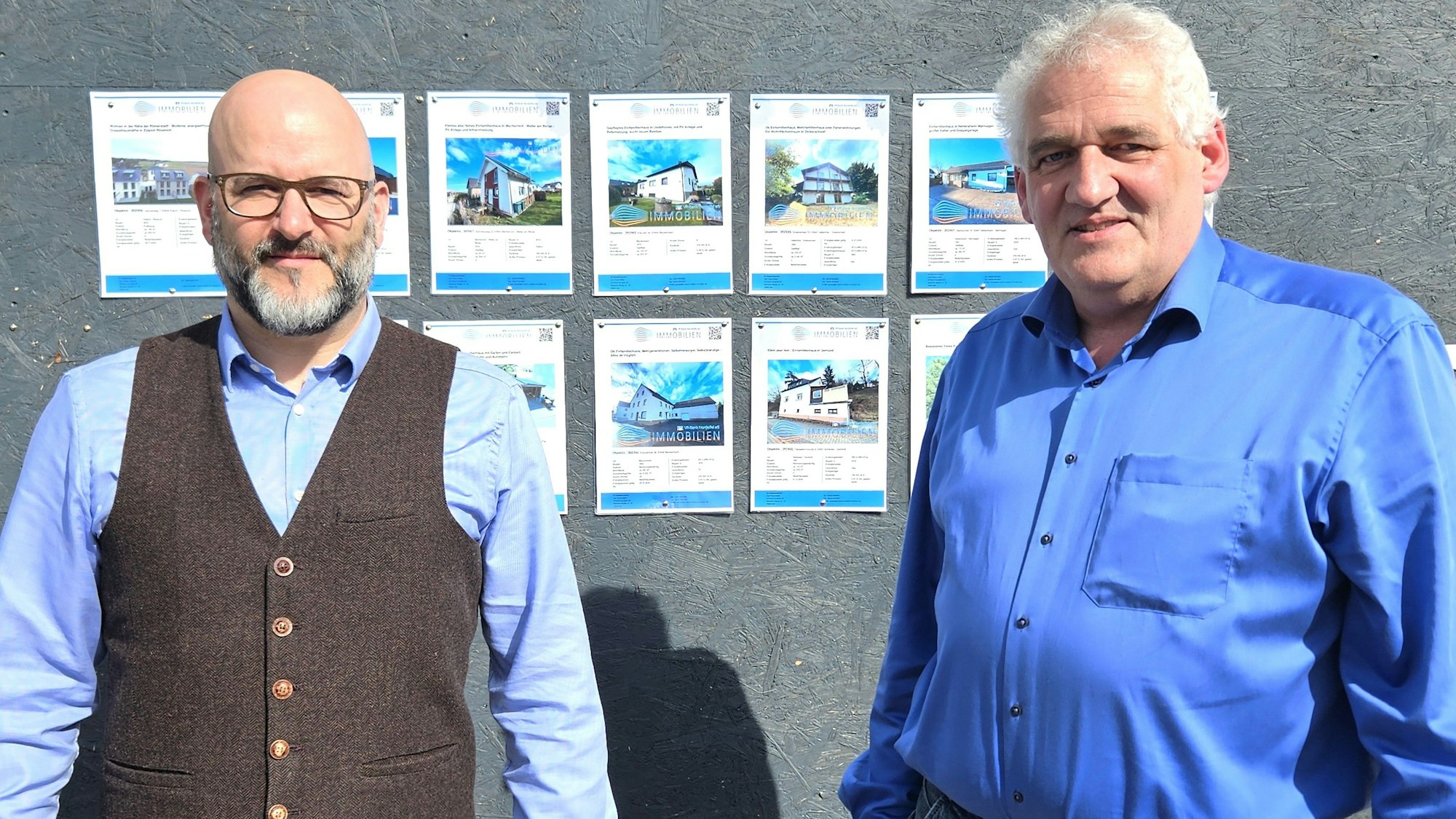 Stefan Lippertz und Hartmut Lackner stehen vor einer Wand mit Immobilienangeboten.