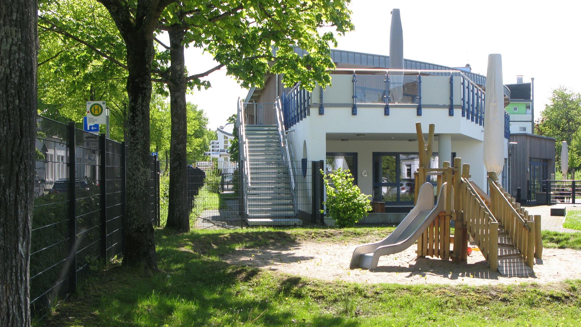 Das zeigt ein zweigeschossiges Kita-Gebäude mit Außentreppe. Das steht in einer Sandkuhle ein Klettertürmchen mit Rutschbahn.