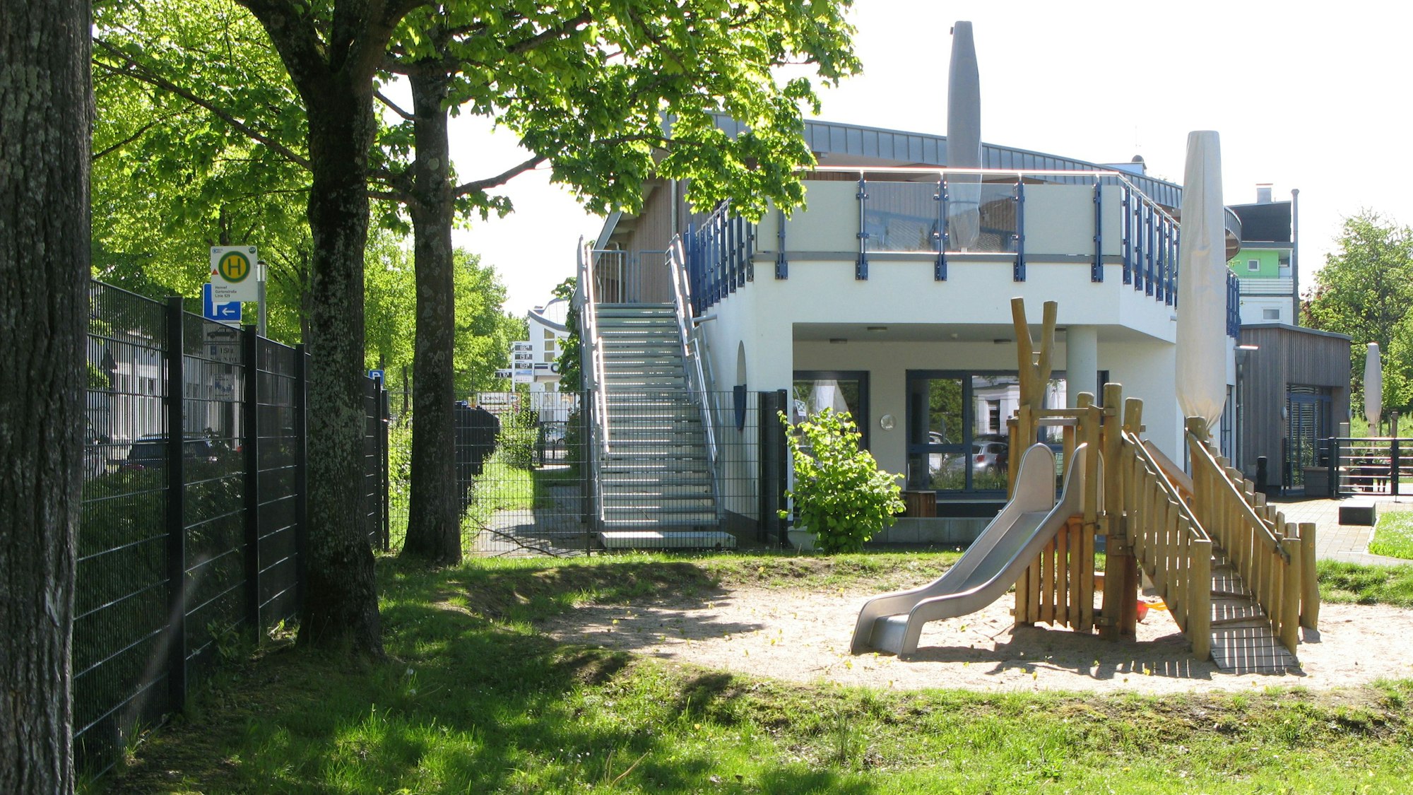 Das zeigt ein zweigeschossiges Kita-Gebäude mit Außentreppe. Das steht in einer Sandkuhle ein Klettertürmchen mit Rutschbahn.