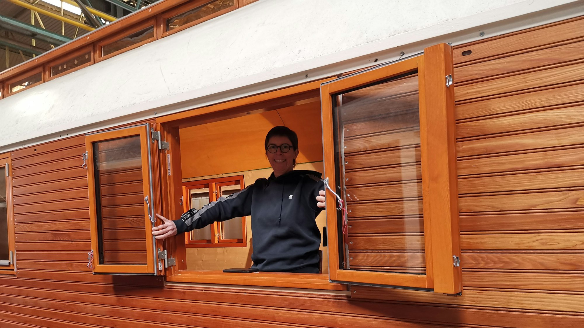 Eine Frau öffnet ein zweiflügeliges Fenster eines hölzernen Zirkuswagens.