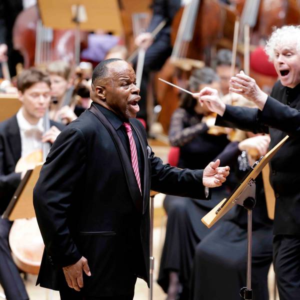 Lester Lynch hat den linken Arm zum Gesang erhoben. Er trägt einen schwarzen Anzug und eine rot-weiß gestreifte Krawatte. Rechts von ihm der Dirigent Simon Rattle, ebenfalls mit schwarzem Anzug. Im Hintergrund sieht man das restliche Orchester verschwommen, sie sitzen an ihren Notenpulten.
