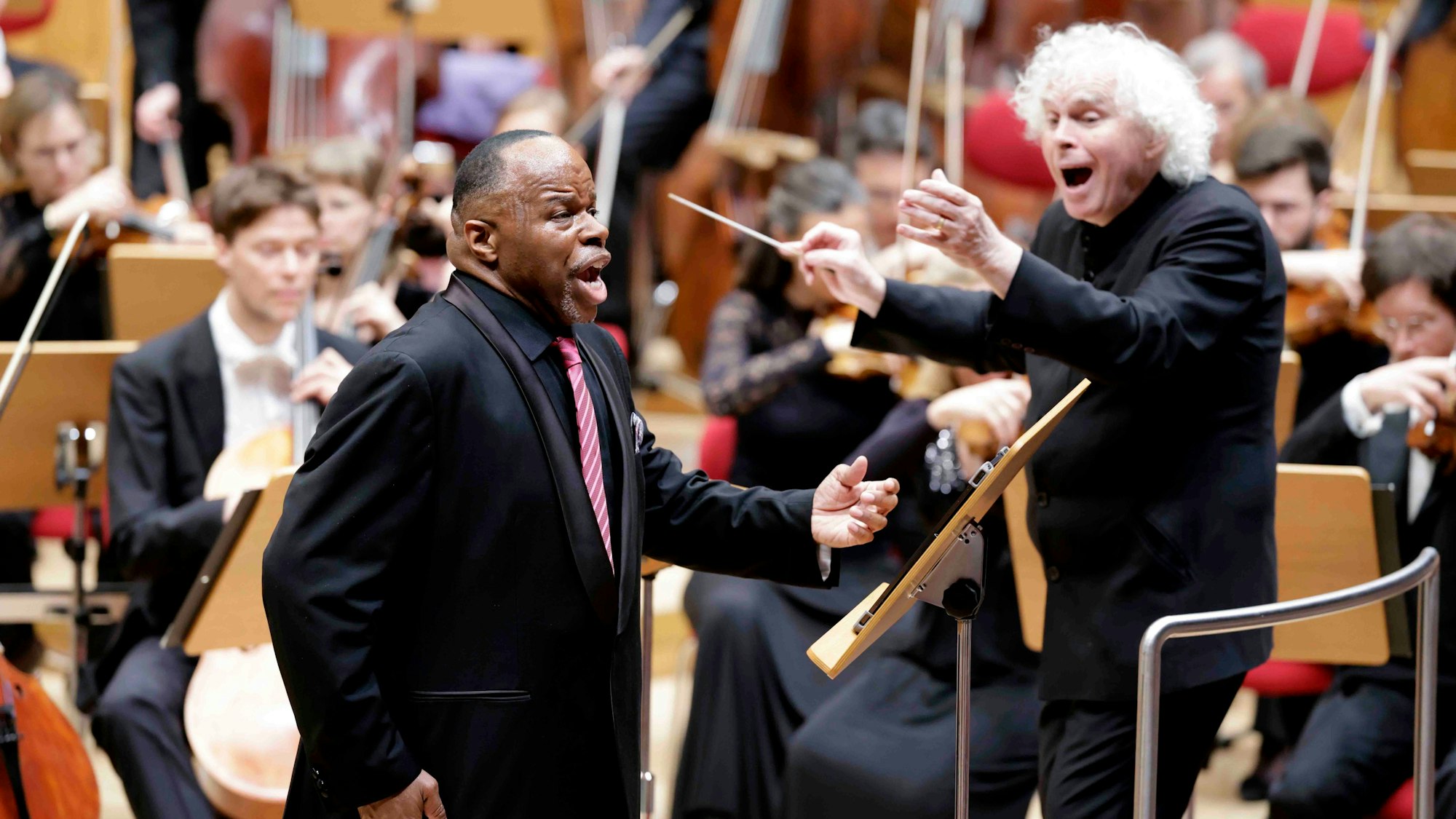 Lester Lynch hat den linken Arm zum Gesang erhoben. Er trägt einen schwarzen Anzug und eine rot-weiß gestreifte Krawatte. Rechts von ihm der Dirigent Simon Rattle, ebenfalls mit schwarzem Anzug. Im Hintergrund sieht man das restliche Orchester verschwommen, sie sitzen an ihren Notenpulten.