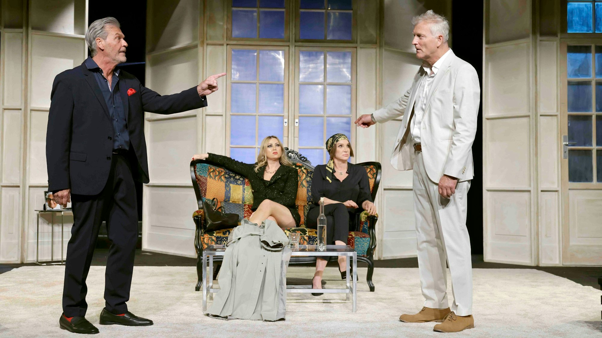 Auf der Bühne stehen die beiden Freunde Bernhard und Philippe. Bernhard zeigt und blickt eindringlich auf seinen Freund. Dieser schaut verdutzt drein und deutet auf die Damen, die im Hintergrund auf einer Couch sitzen und auf Philippe zurückschauen.