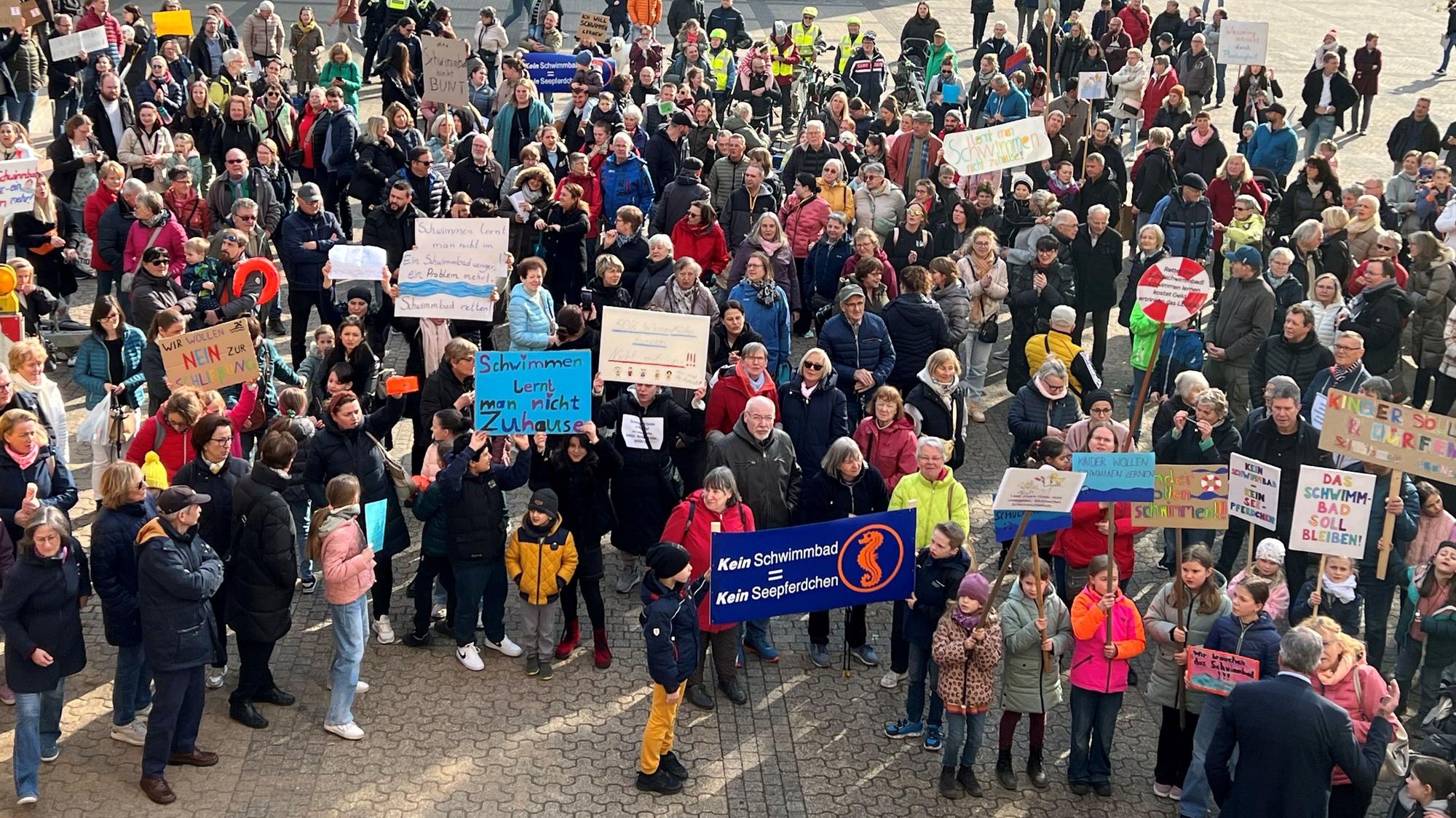 Auf dem Foto sind protestierende Menschen zu sehen. Sie halten Plakate gegen die Schließung des Schulschwimmbads hoch.