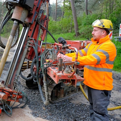 Ein Arbeiter bedient eine Bohrraupe, die auf einem Waldweg steht. Er trägt eine Jacke in Signalfarbe und einen Schutzhelm.