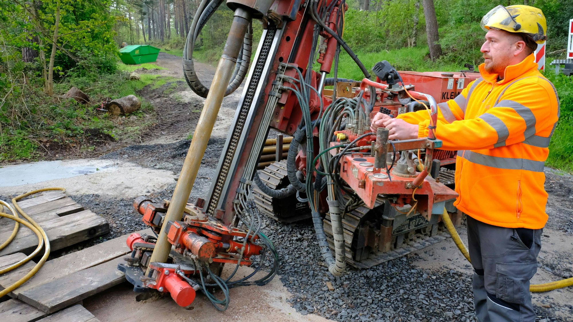 Ein Arbeiter bedient eine Bohrraupe, die auf einem Waldweg steht. Er trägt eine Jacke in Signalfarbe und einen Schutzhelm.
