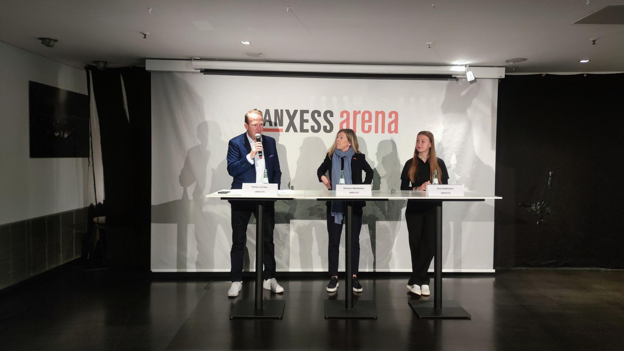 Ein Mann und zwei Frauen stehen vor einem Plakat mit der Aufschrift „Lanxess Arena“