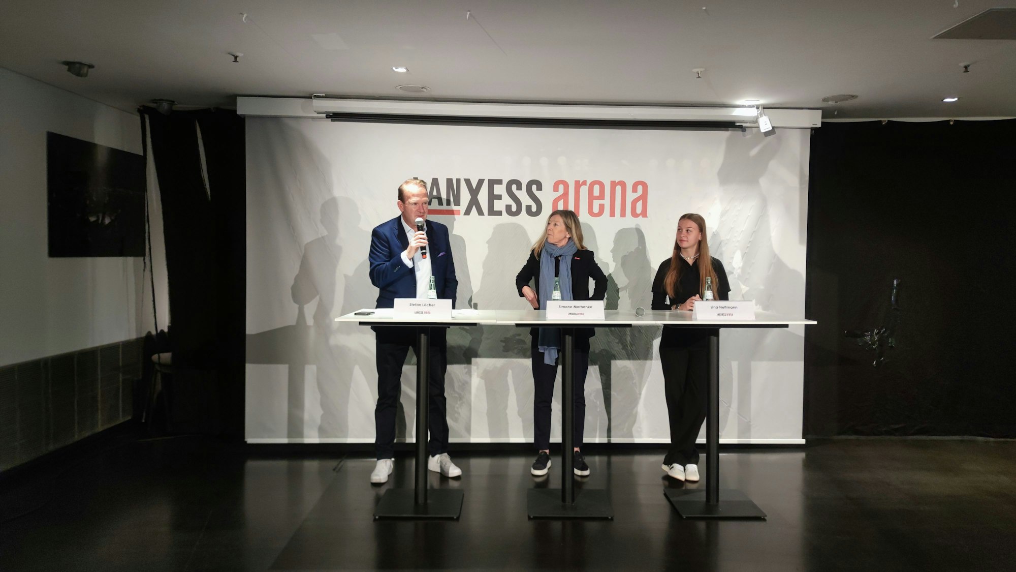 Ein Mann und zwei Frauen stehen vor einem Plakat mit der Aufschrift „Lanxess Arena“