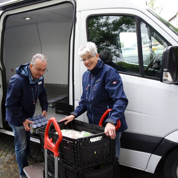 Das Foto zeigt eine Frau und einen Mann, die Lebensmittel aus einem Auto ausladen.
