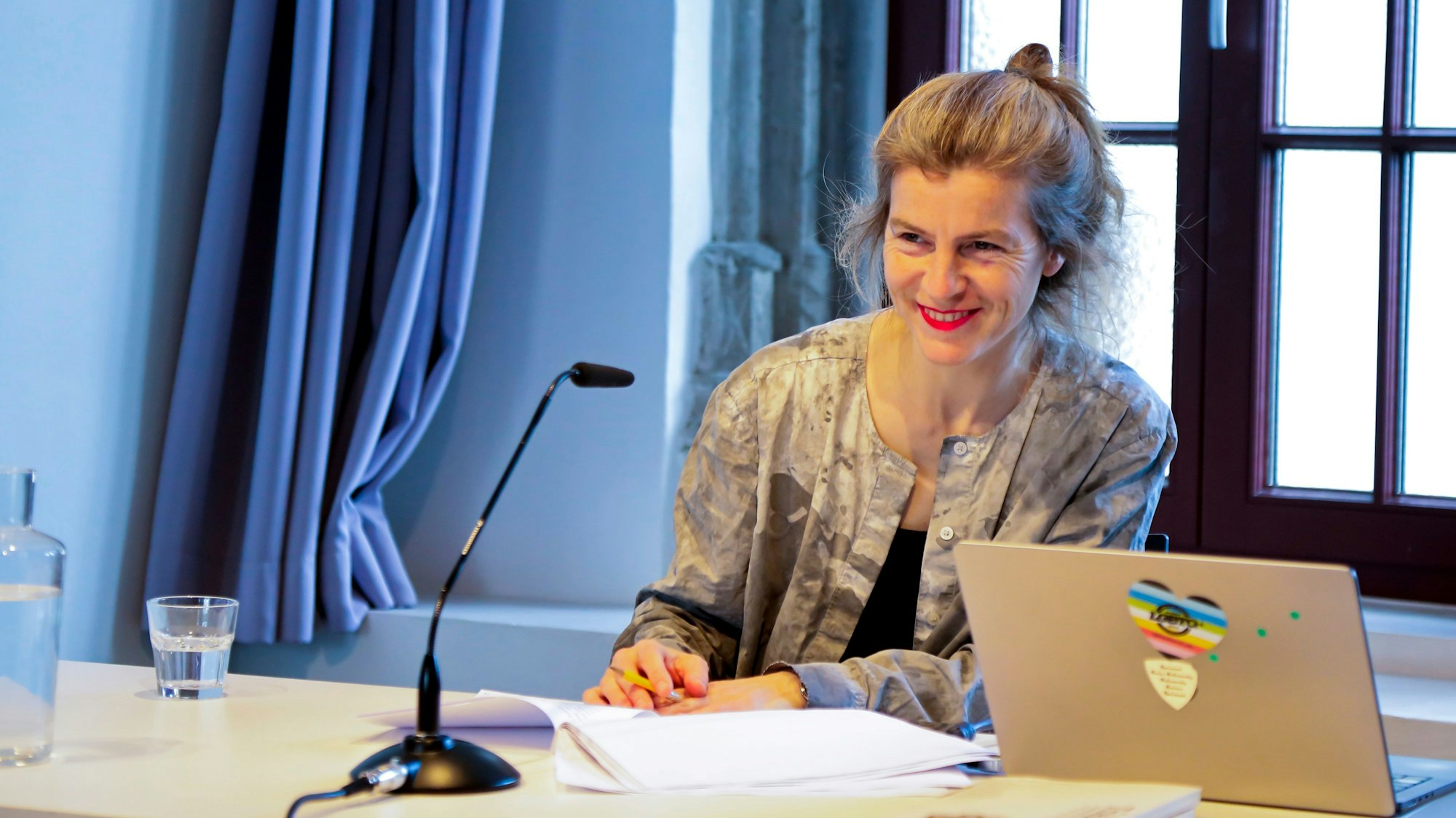 Anne Cotten sitzt an einem Schreibtisch, vor sich ein Stapel Papiere, ein Laptop und einem Mikrofon. Sie hat ihre Haare hinten zu einem Topf zusammengebunden.