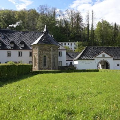 Eine Kapelle in einer Wiese, dahinter ein Hotel und ein historischer Küchenhof.