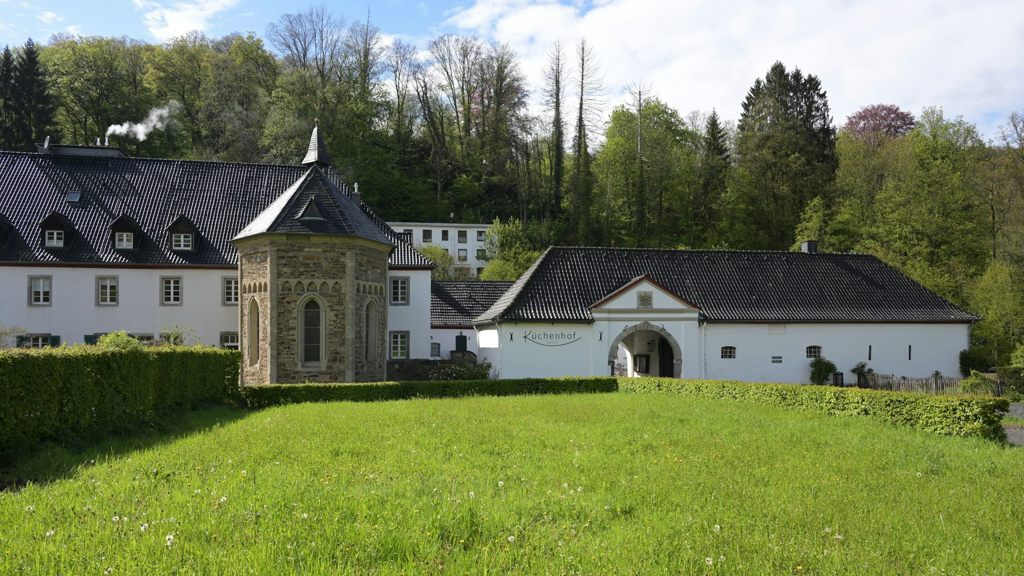 Eine Kapelle in einer Wiese, dahinter ein Hotel und ein historischer Küchenhof.