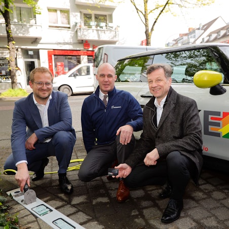 Drei Männer knien vor einem E-Auto, das an einer neuen Ladestation geladen wird.