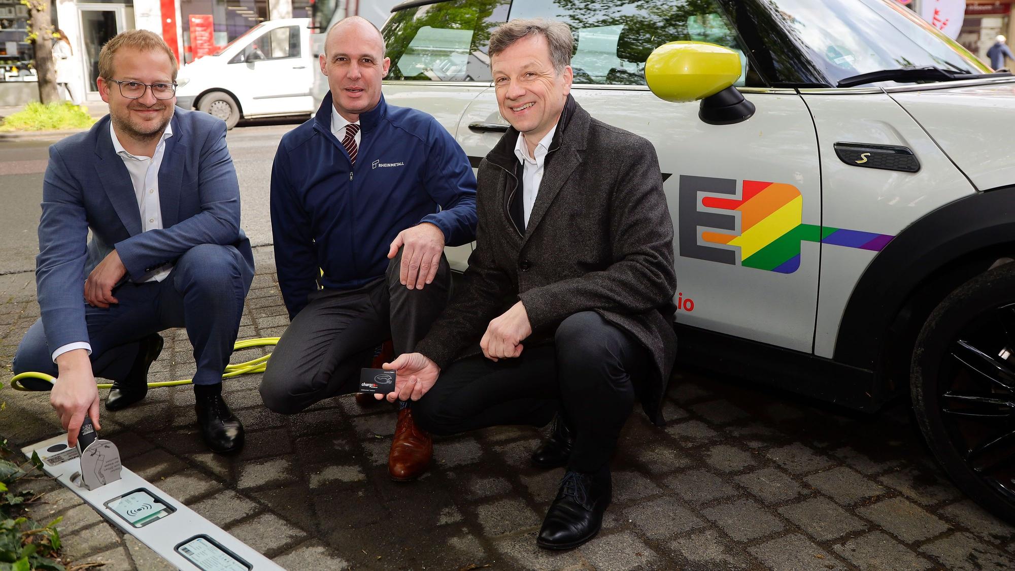 Drei Männer knien vor einem E-Auto, das an einer neuen Ladestation geladen wird.