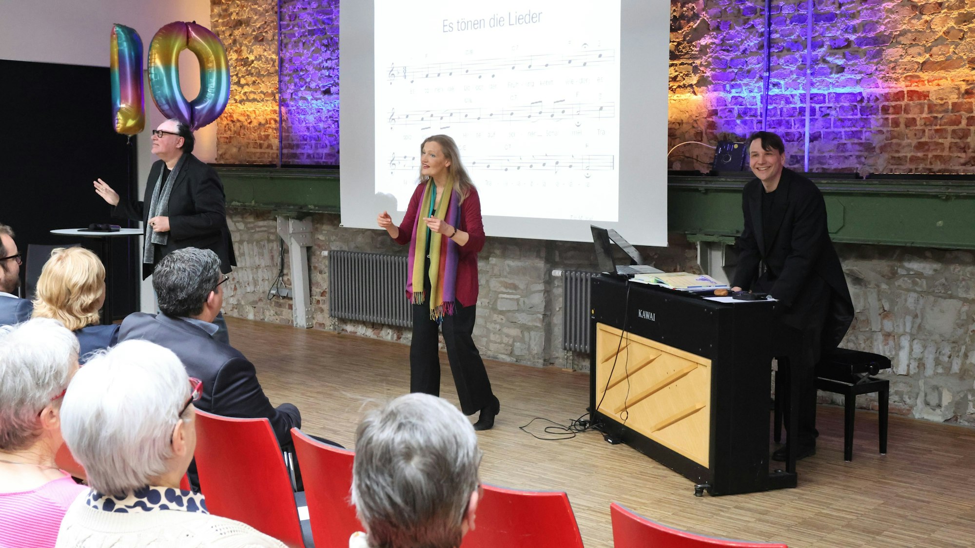 Eine Dirigentin steht vor einer Leinwand mit Noten und leitet einen Seniorenchor an.