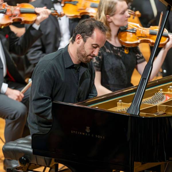 Auf dem Bild ist Igor Levit zu sehen, wie er beim Spielen konzentriert auf seinen Flügel schaut. Er trägt ein schwarzes Hemd. Im Hintergrund weitere Musikerinnen und Musiker.