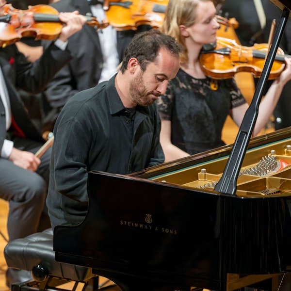Auf dem Bild ist Igor Levit zu sehen, wie er beim Spielen konzentriert auf seinen Flügel schaut. Er trägt ein schwarzes Hemd. Im Hintergrund weitere Musikerinnen und Musiker.