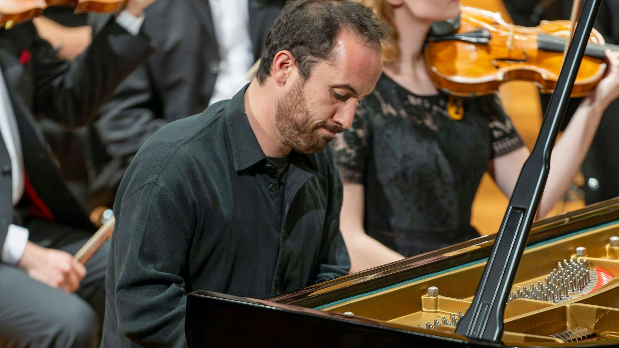 Auf dem Bild ist Igor Levit zu sehen, wie er beim Spielen konzentriert auf seinen Flügel schaut. Er trägt ein schwarzes Hemd. Im Hintergrund weitere Musikerinnen und Musiker.