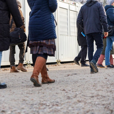 Die Beine von Menschen laufen über einen geschotterten Platz, im Hintergrund sind Wohncontainer für Geflüchtete zu sehen.