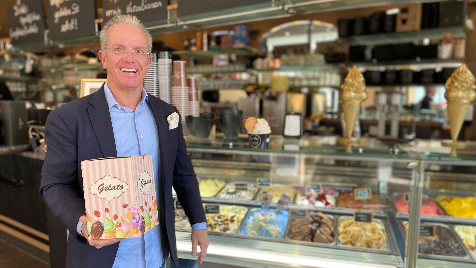 Andreas Siebrecht trägt einen Anzug. Vor der Eisauslage präsentiert er einen Pappkarton mit der Aufschrift „Gelato“.
