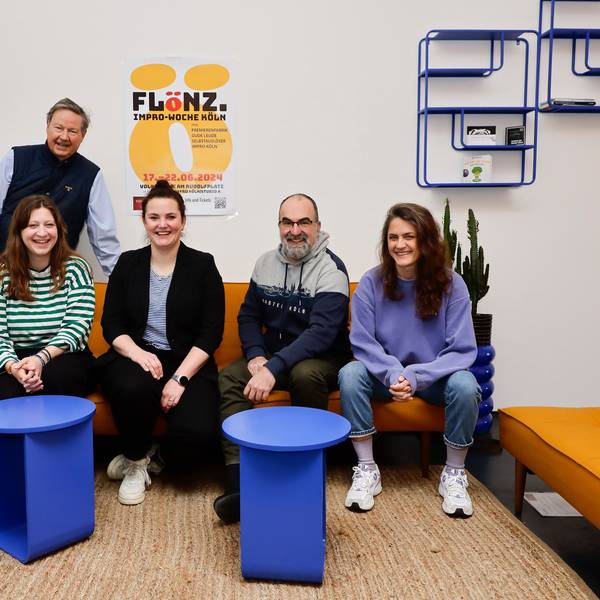 Zwei Männer und drei Frauen posieren rund um ein gelbes Sofa für ein Foto. Hinter ihnen ist ein Plakat zu sehen.