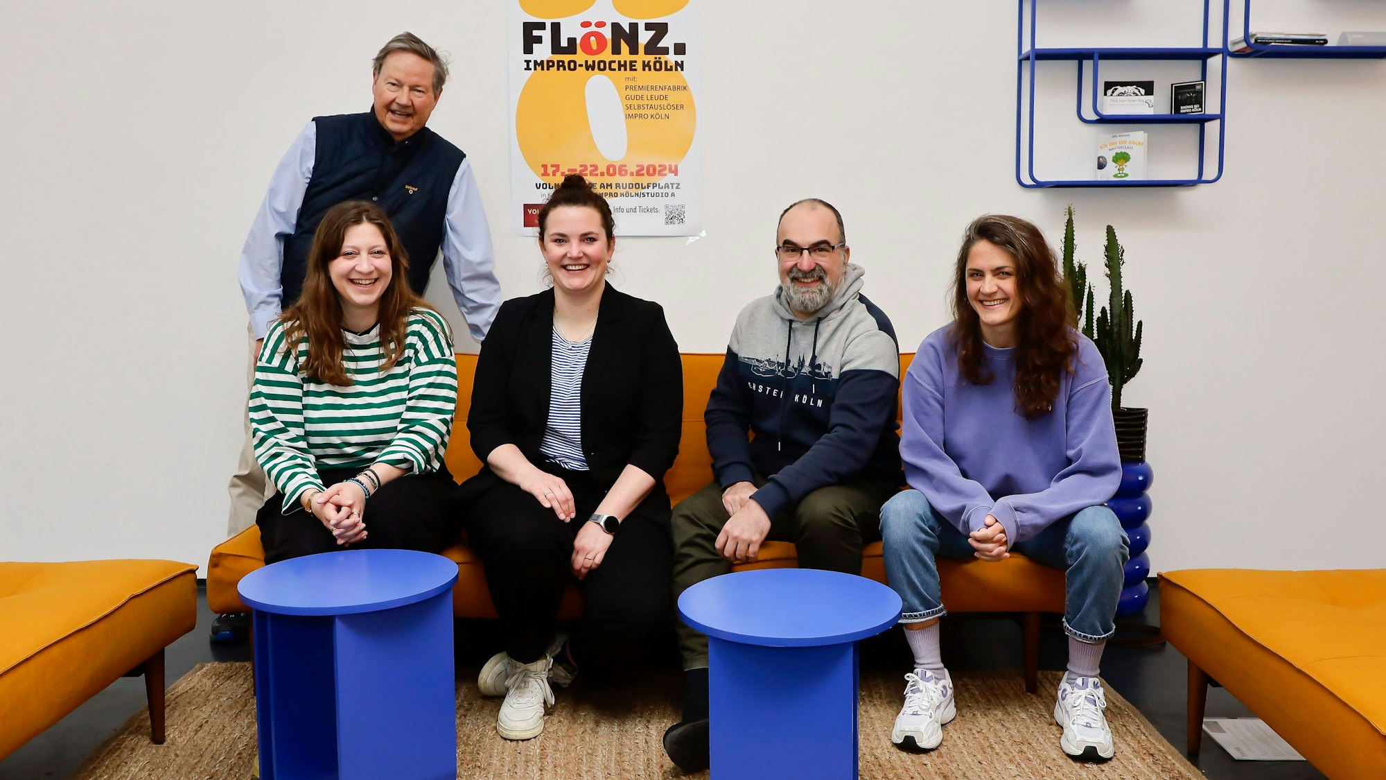 Zwei Männer und drei Frauen posieren rund um ein gelbes Sofa für ein Foto. Hinter ihnen ist ein Plakat zu sehen.