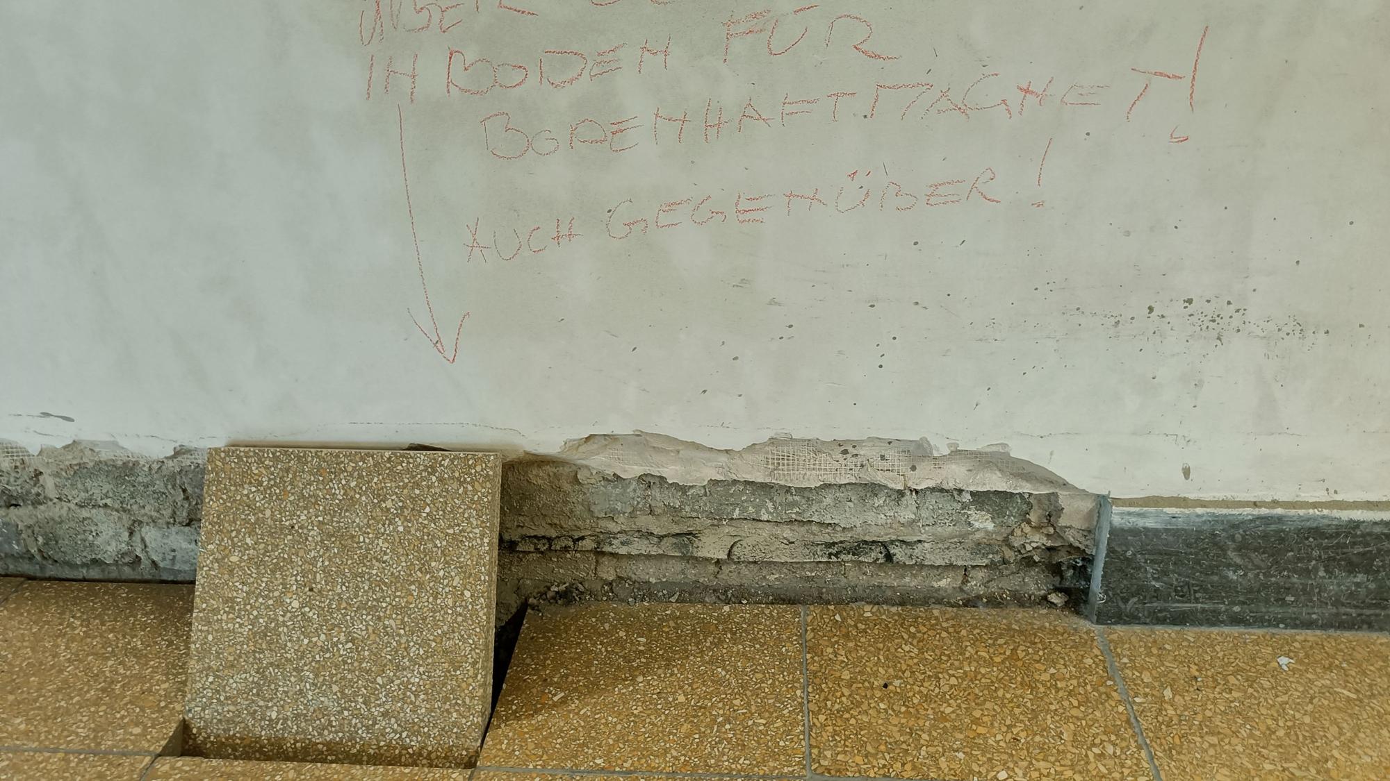 Eine Bodenfiese ist aus dem Boden gelöst und lehnt an einer maroden Wand.
