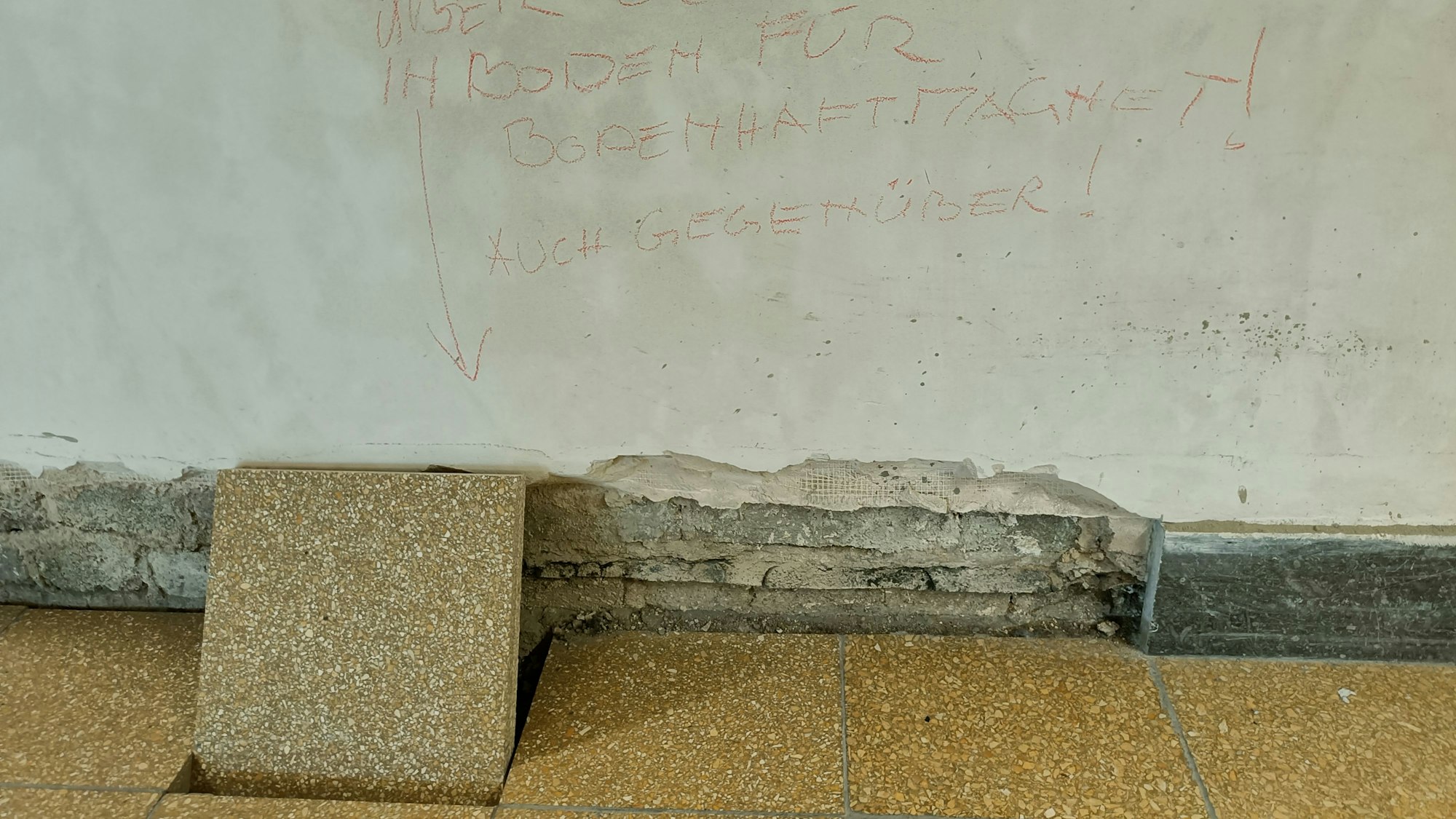 Eine Bodenfiese ist aus dem Boden gelöst und lehnt an einer maroden Wand.