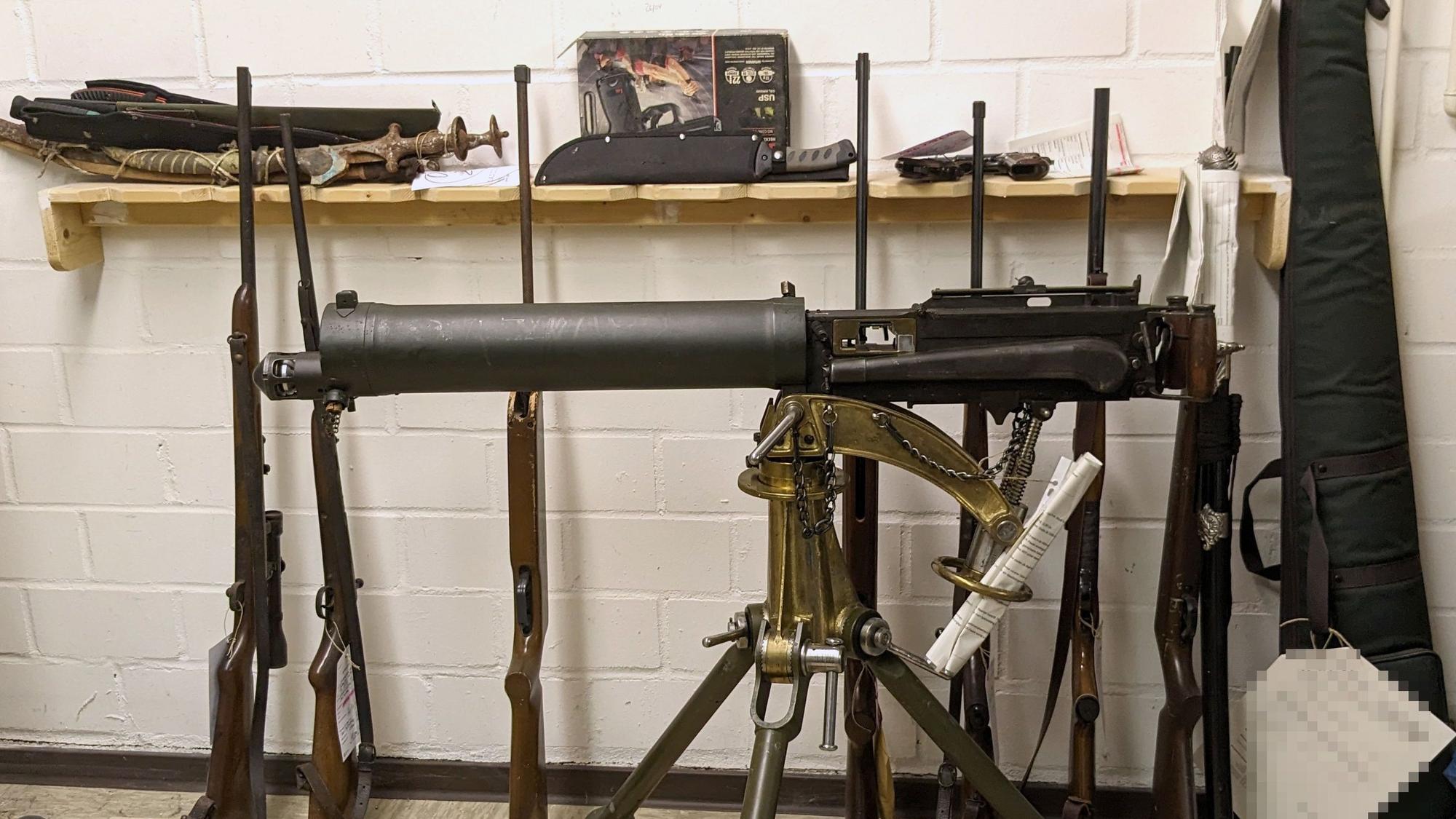 Ein altes Maschinengewehr ist auf einem Dreibein montiert. Dahinter lehnen diverse Waffen an der Wand.