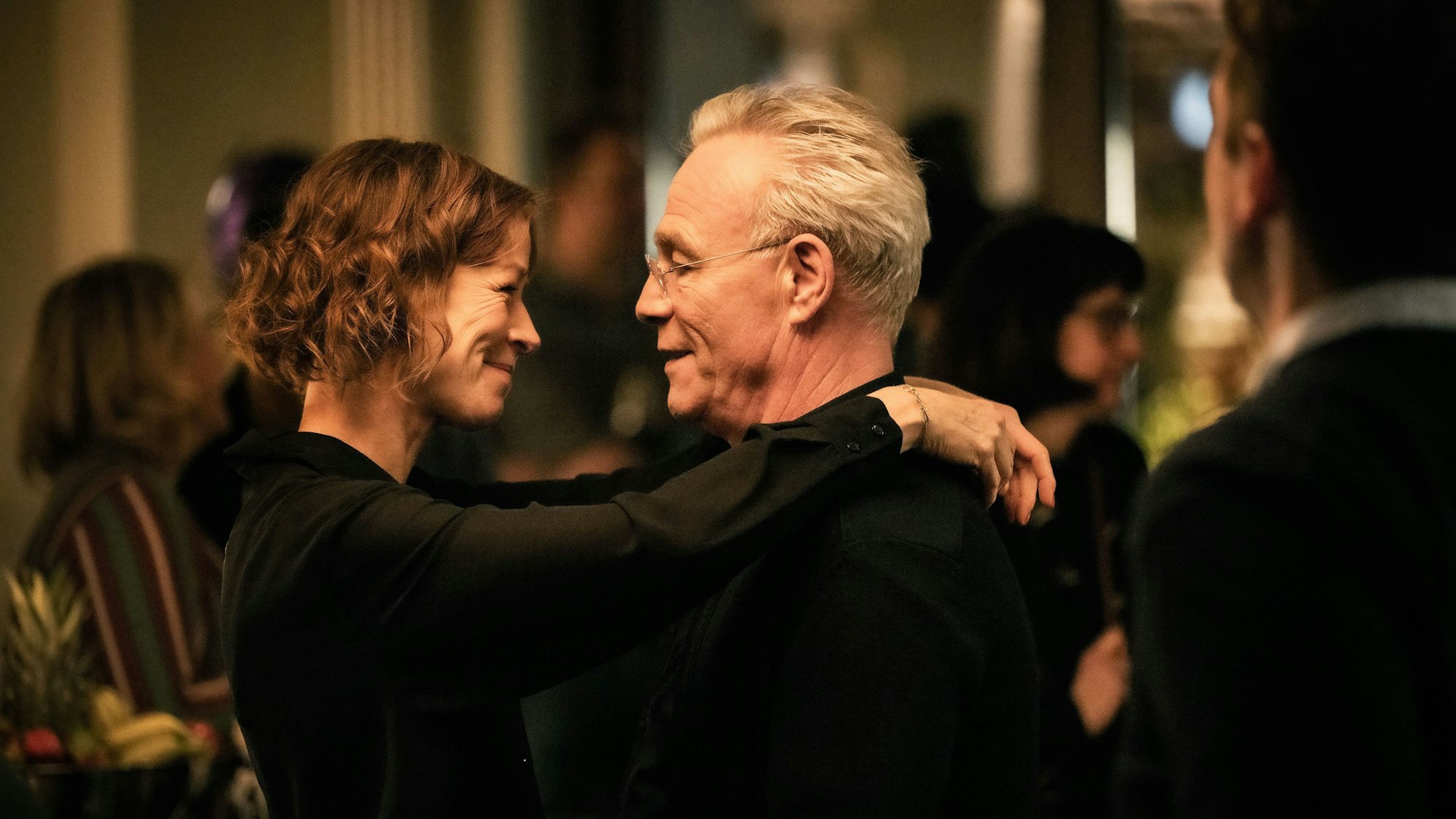 Auf dem Bild liegen sich Kommissar Ballauf und seine Freundin Nicola Koch in den Armen, um sie herum andere Menschen. Das Pärchen wirkt vertraut, lächelt sich an.