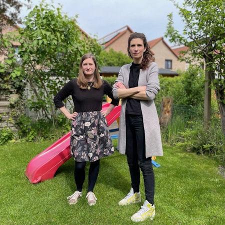 Zwei Frauen in einem Garten; im Hintergrund steht eine bunte Kinderrutsche aus Kunststoff.
