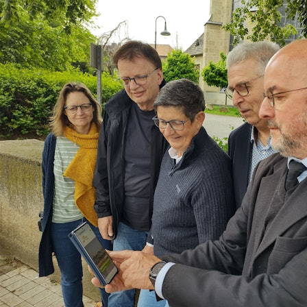Werner Jacobs hat auf dem Handy die Homepage des Projekts „Lebendiger Friedhof“ aufgerufen, die anderen Projekt-Mitwirkenden schauen mit ihm darauf.