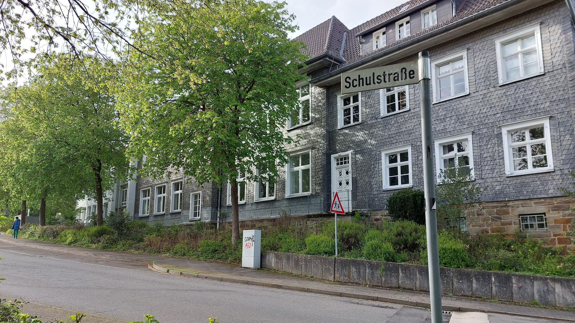 Ein mit grauem Schiefer verkleidetes Schulgebäude mit weißen Fensterrahmen. Vor dem Gebäude verläuft eine ansteigende Straße; auf dem Straßenschild steht Schulstraße