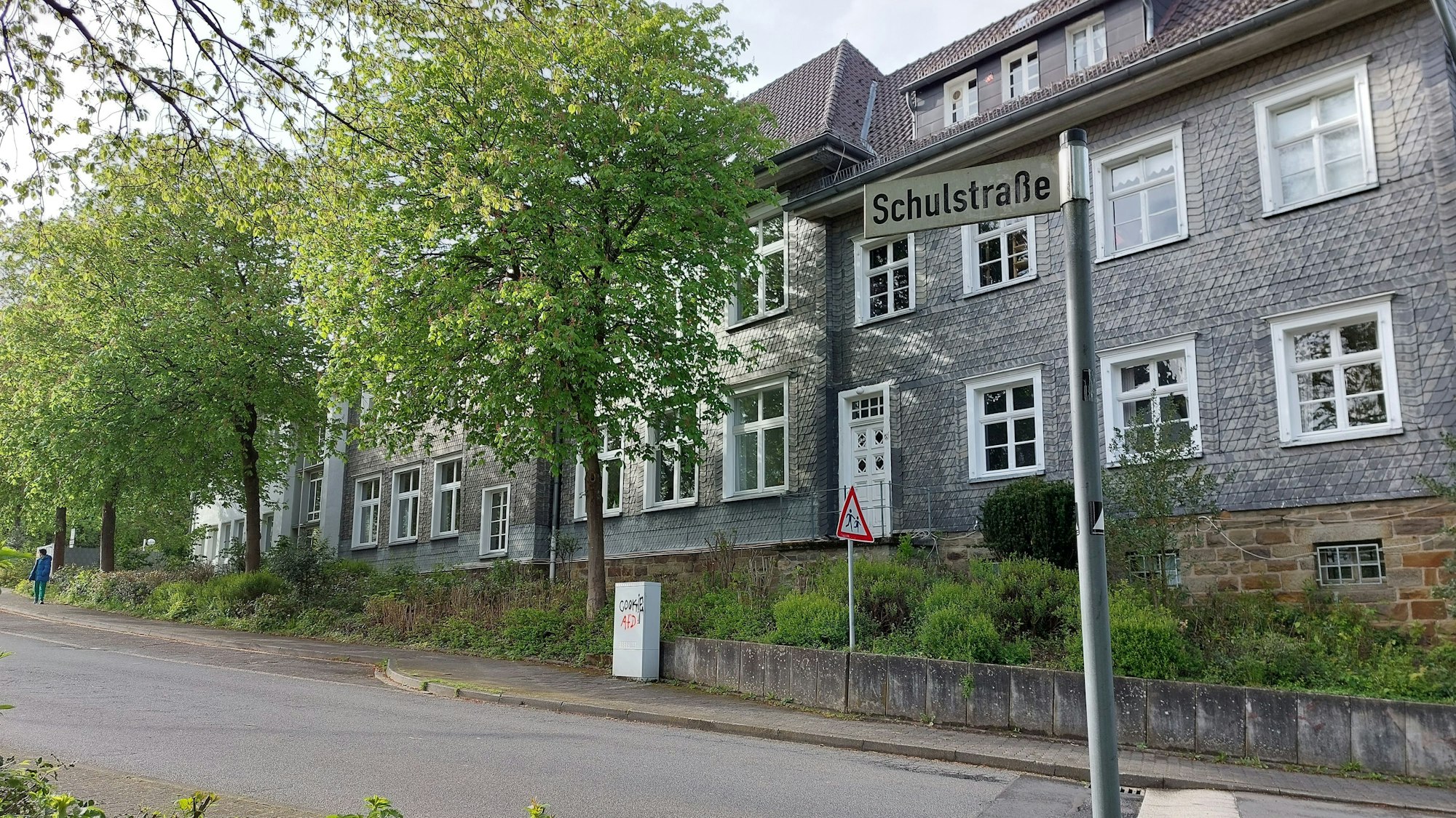 Ein mit grauem Schiefer verkleidetes Schulgebäude mit weißen Fensterrahmen. Vor dem Gebäude verläuft eine ansteigende Straße; auf dem Straßenschild steht Schulstraße