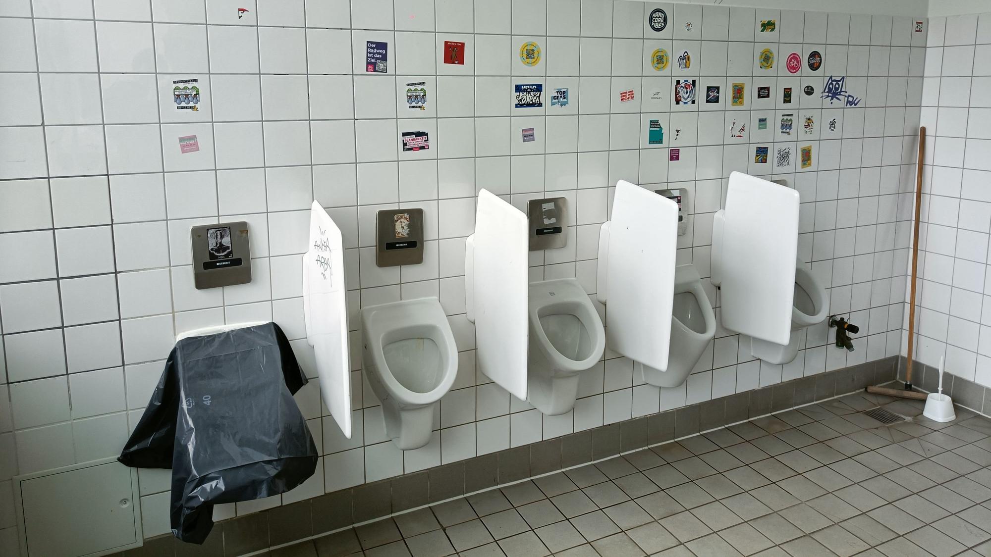 Das linke von fünf Pissoirs ist mit einer Plane abgedeckt. Die Wand ist voll mit Aufklebern.