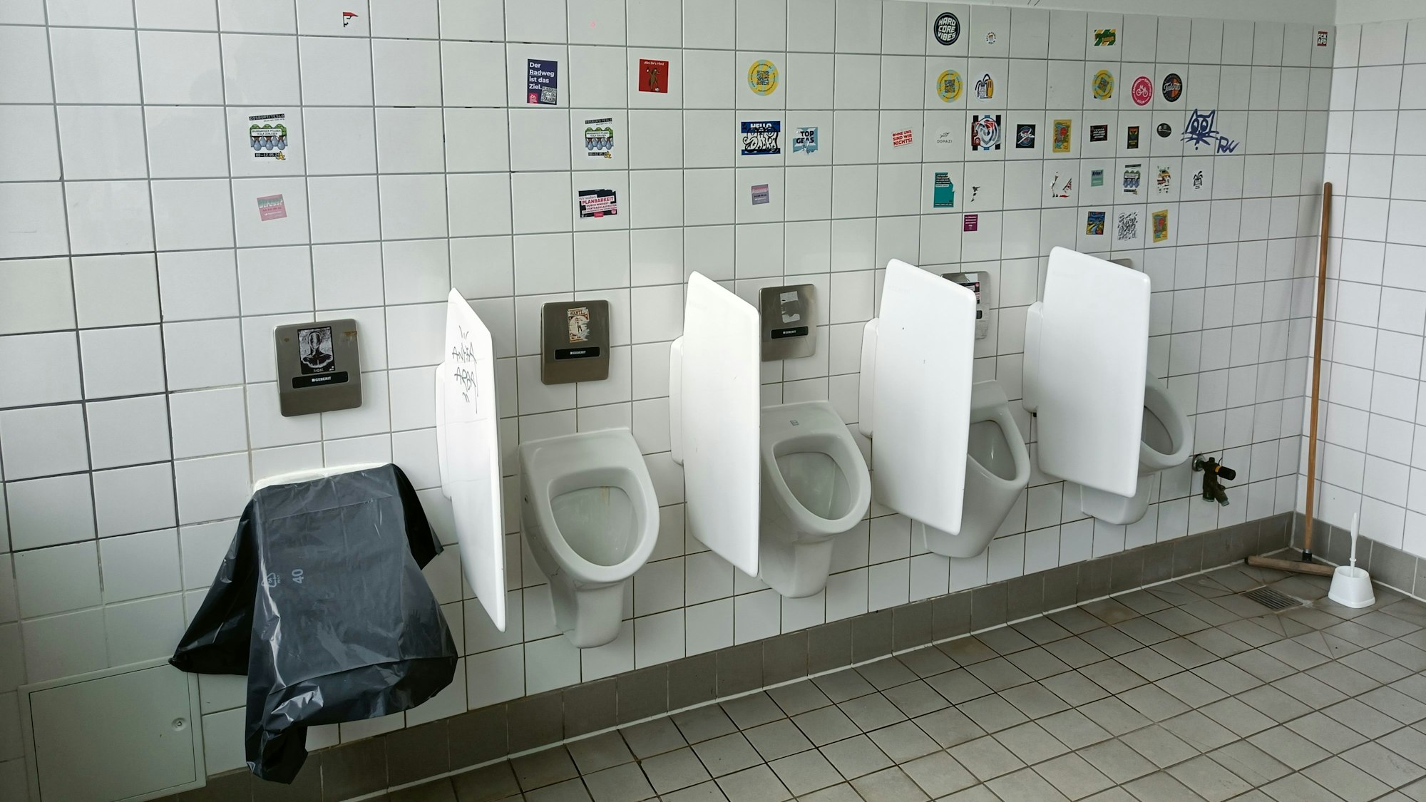 Das linke von fünf Pissoirs ist mit einer Plane abgedeckt. Die Wand ist voll mit Aufklebern.