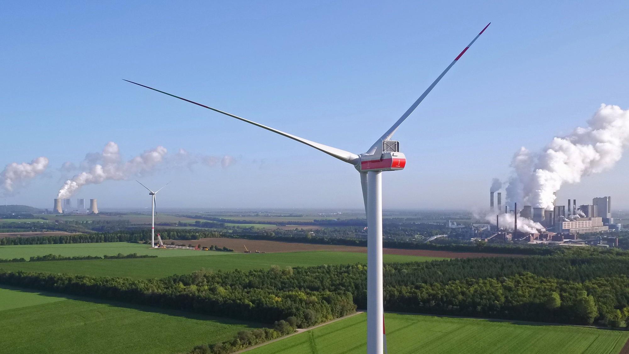 Zwei Windräder, im Hintergrund sind Kraftwerke zu sehen.